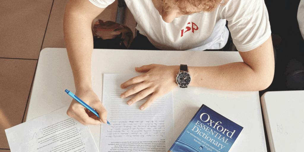 A person wearing a white tshirt is writing an essay using a pen. They have a copy of the Oxford English Dictionary next to them.
