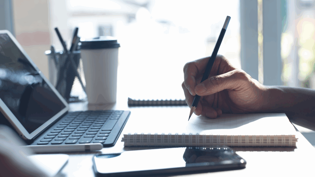 A person writing in a notebook with a computer and a cup of coffee in front of them.