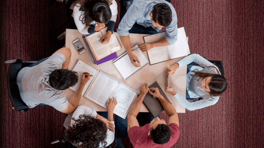 A shot from above of 6 students around a table with books, notebooks, phones and textbooks